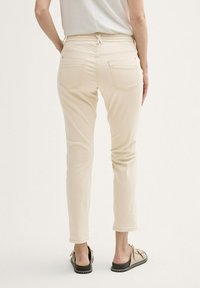 Pantaloni tapered beige con una texture liscia, dotati di due tasche posteriori e vita media. Abbinati a scarpe beige slip-on.