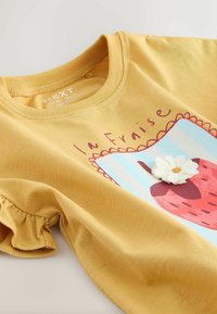 Tricou galben din bumbac cu un desen cu căpșuni și un accent de floare albă. Detaliu cu mâneci cu volănașe și design cu decolteu rotund.