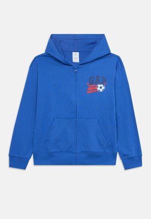 Sweat à capuche bleu zippé avec poches avant, capuche et logo « GAP » avec un ballon de football et des rayures rouges sur la poitrine.