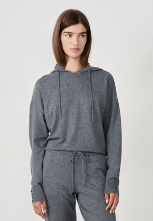 Young woman wearing a grey knit hooded sweater and matching knit pants with drawstring and buttoned cuffs, standing against a plain light background.