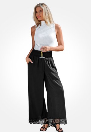 Schwarze Hosen mit weitem Bein, Spitzenbesatz am Saum und elastischem Bund, kombiniert mit einem enganliegenden weißen ärmellosen Oberteil. Eine klare Cocktailglas wird gehalten.