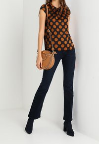 Kvinna som bär en svart och brun prickig blus, mörkblå jeans, svarta stövlar och bär en rund brun axelväska.