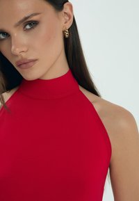 Czerwona bluzka na ramiączkach z gładkiej, elastycznej tkaniny. Posiada wysoki kołnierz i odkryte ramiona, w połączeniu ze złotymi kolczykami w kształcie kół.