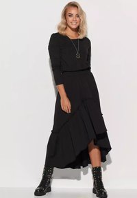 Uśmiechnięta kobieta z blond włosami, ubrana w czarną bluzkę z długim rękawem, asymetryczną czarną spódnicę, naszyjnik z zawieszką oraz czarne buty ze ćwiekami.