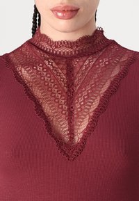 Burgundy topp med ribbad textur; har en spetskragdesign i mörk burgundy som skapar en V-form med intrikata detaljer.
