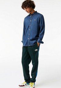 Granatowa koszula jeansowa na guziki, zestawiona z zielonymi sportowymi spodniami z niebieskimi bocznymi paskami i haftowanym logo. Białe trampki z akcentami.