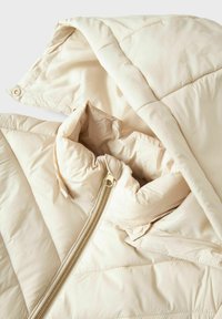 Chaqueta acolchada beige con capucha, que presenta un diseño acolchado y una cremallera dorada. El interior incluye un forro suave y un cierre de botón.