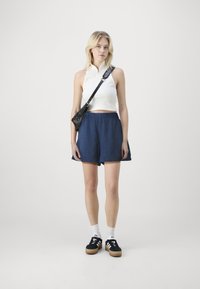 Baltas be rankovių marškinėlis su aukštu apykakle, derinamas su mėlynomis lino šortais ir juodais sportbačiais. Komplektą užbaigia mažas juodas per petį nešamas krepšys.