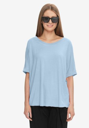 Kobieta nosząca oversize jasnoniebieską bluzkę z krótkim rękawem i duże czarne okulary przeciwsłoneczne, uśmiechająca się na jednolitym białym tle.