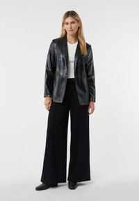 Czarna skórzana marynarka o błyszczącej fakturze, z klapami i dwoma przednimi kieszeniami, zestawiona z szerokimi czarnymi spodniami oraz białą bluzką.