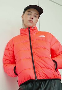 Giacca arancione gonfia con colletto alto e zip frontale. Presenta sezioni trapuntate e un logo sul lato sinistro del petto. Cappellino nero indossato.