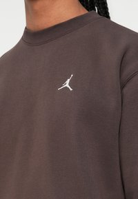Acercamiento de una persona que lleva puesta una sudadera marrón con un pequeño logo Jumpman bordado en el pecho.