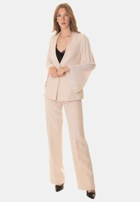 Abito beige su misura con blazer strutturato e maniche plissettate, abbinato a un top nero e pantaloni dritti, indossato con tacchi neri.