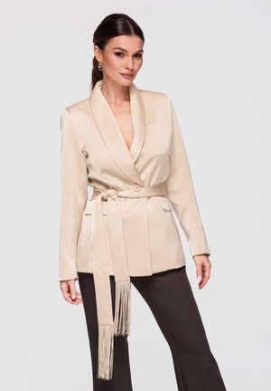 Donna in piedi che indossa un blazer beige con cintura e cravatta con frange e pantaloni neri su uno sfondo chiaro e uniforme.