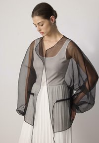 Crni prozirni kimono s labavim rukavima, kontrastnim obrubom i dva prednja džepa iznad sive majice i bijele plisirane suknje. Lagana tekstura.