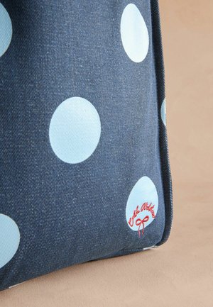 Denim tas met grote lichtblauwe stippen, voorzien van een rood geborduurd logo op de rand, tegen een zachte beige achtergrond.
