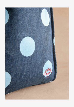 Sac en denim avec de grands pois bleu clair, orné d'un logo brodé rouge sur le bord, le tout sur un fond beige doux.