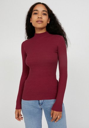 Bordeauxroter, gerippter Langarmpullover mit hohem Kragen, figurbetonter Silhouette und weicher Textur, kombiniert mit hellblauen Jeans.