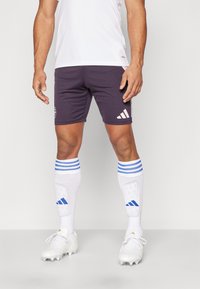 Camiseta deportiva blanca, pantalones cortos morados oscuros con logo y tres rayas blancas, calcetas blancas con rayas azules y zapatillas blancas con tacos.