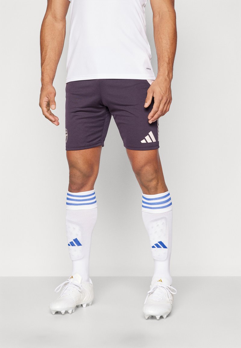 Camiseta deportiva blanca, pantalones cortos morados oscuros con logo y tres rayas blancas, calcetas blancas con rayas azules y zapatillas blancas con tacos.
