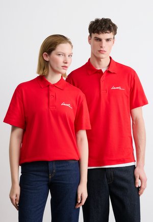 Mladi moški in ženska, oblečena v ujemajoče se rdeče polo majice z belim logotipom "Lacoste", kombinirane s temno modrimi jeans hlačami, stojita pred belo ravno steno.