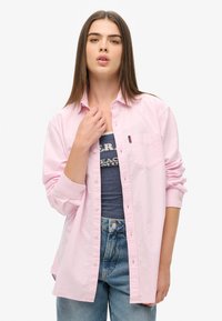 Jasnoróżowa koszula zapinana na guziki z kieszonką na piersi, z podwiniętymi rękawami i luźnym krojem, zestawiona z ciemnoniebieskim topem na ramiączkach i jasnymi dżinsami.