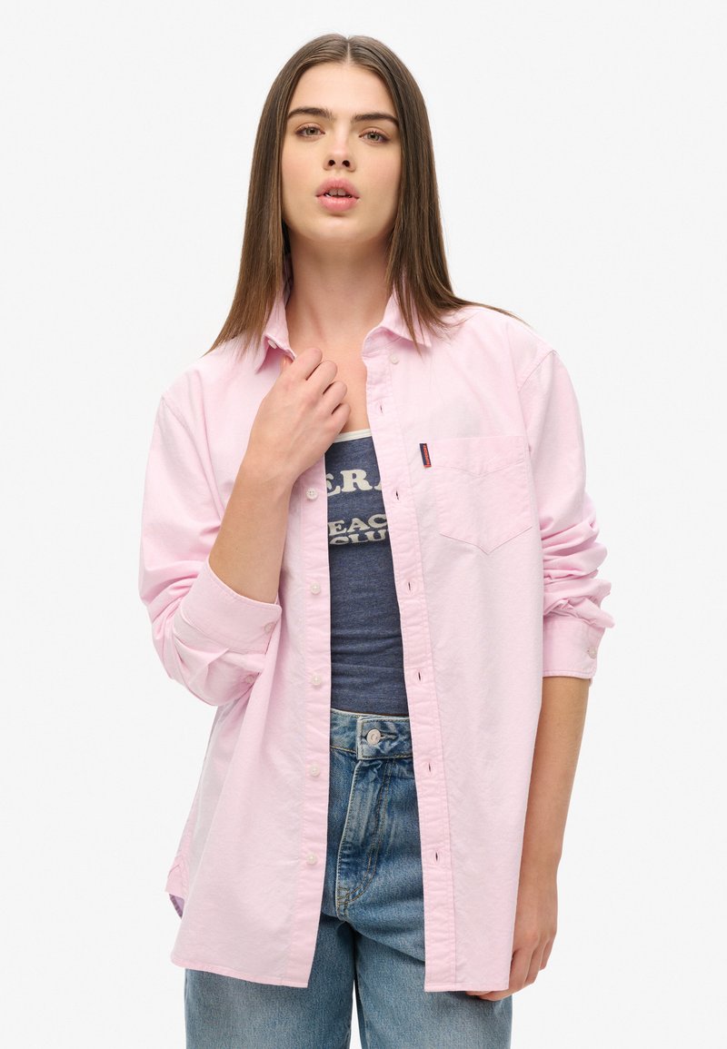 Jasnoróżowa koszula zapinana na guziki z kieszonką na piersi, z podwiniętymi rękawami i luźnym krojem, zestawiona z ciemnoniebieskim topem na ramiączkach i jasnymi dżinsami.