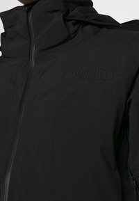 Veste noire à col haut, fabriquée en matériau lisse et résistant à l'eau. Dispose d'une fermeture éclair à l'avant et d'un logo en relief sur la poitrine.