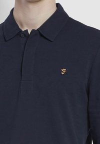 Námořnická polo košile vyrobená z texturované látky, s límečkem na dva knoflíky a malým zlatým logem na levé části hrudi.