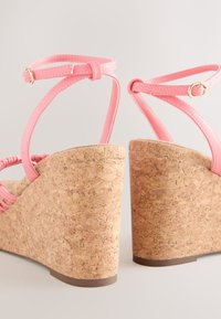 Lyserøde kilehælssandaler med korkplatform, der har tynde ankelsnore og en tekstureret overflade. Guldhardwaredetaljer på stroplukninger.