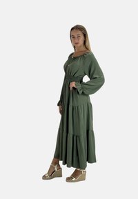 Lang grøn kjole med rynket talje, brede ærmer og lagdelt nederdel. Kombineret med sølv kilehælssandaler. Glat, tekstureret stof.