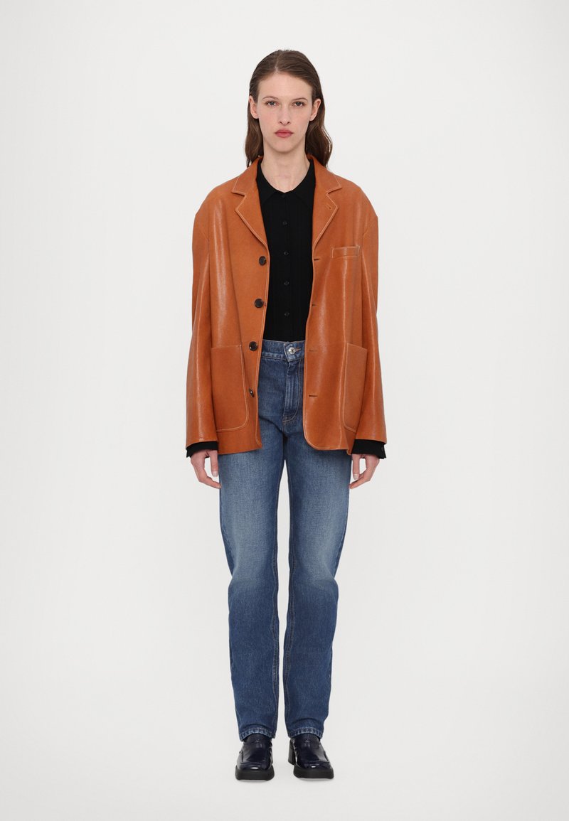 Veste en cuir marron avec une coupe décontractée, col cranté et boutons. Associée à un jean en denim bleu et une chemise noire, dotée de poches.