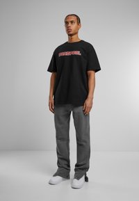 Black cotton T-shirt with "DEADPOOL" logo in red, paired with grey pants and white sneakers. Model stands against a neutral background.
