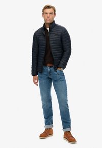 Navy puffer jacket, zippered, textured fabric. Dark brown sweater underneath. Light blue jeans, rolled cuffs. Brown boots with laces.