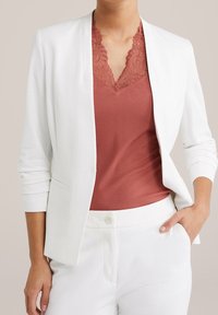 Valkoinen istuva bleiseri ruosteenvärisen, pitsireunuksellisen ribbineuletopin päällä. Malli pitää kädet taskuissa, esitellen tyylikästä muotoilua ja räätälöityä istuvuutta.