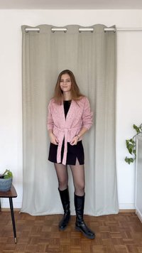 Gaisni rozā teksturēta kardigāna josta ap melnu kleitu, kopā ar augstiem melniem zābakiem. Neitrāla aizkars un augs fona attēlā.