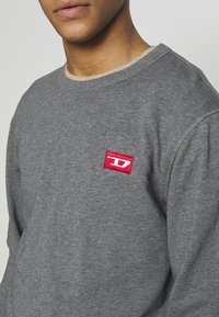 Gros plan d'une personne portant un sweat-shirt gris avec un petit patch logo rouge et blanc sur la partie gauche de la poitrine.