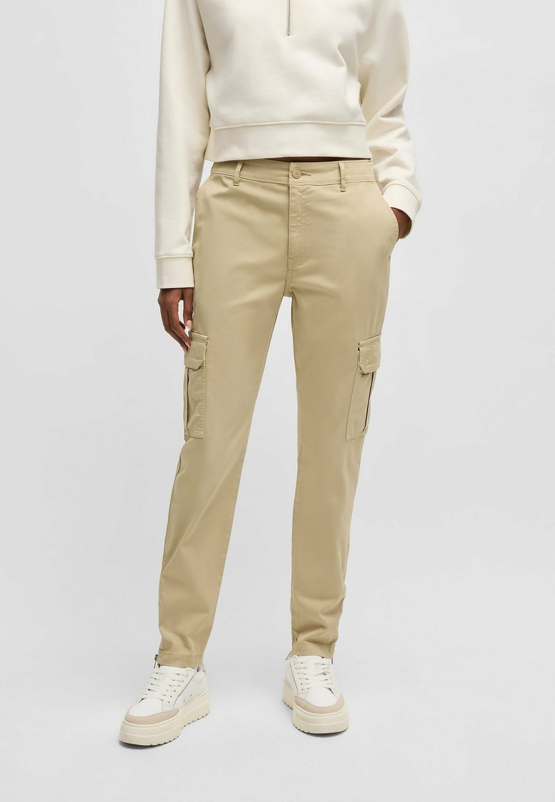 Personne portant un pantalon cargo beige, des baskets à plateforme blanches et un haut à manches longues crème avec une fermeture éclair à l'avant.