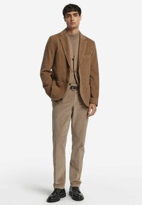 Brauner Cordblazer mit passender Hose, kombiniert mit einem gestrickten beigen Pullover. Merkmale sind ein strukturiertes Design und schwarze Lederhalbschuhe.