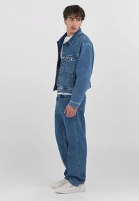 Modra denim jakna z gumbnimi zapiranji, dvema žepoma na prsih in sproščenim krojem, v kombinaciji z ujemajočimi se širokimi temnimi jeans hlačami in belimi supergami.