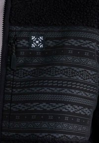 Chaqueta de forro polar negra con patrones geométricos en gris oscuro y un logo cuadrado bordado en blanco encima de un bolsillo con cremallera.