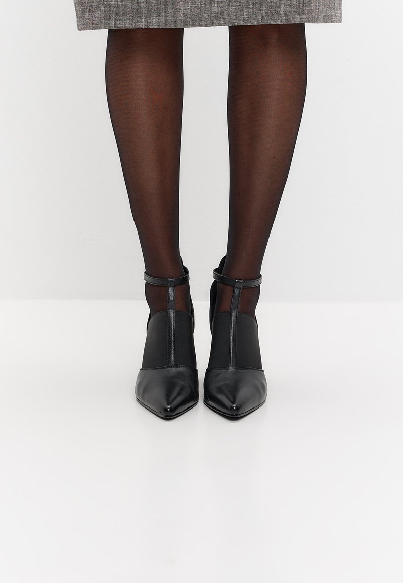 Piernas con medias negras transparentes que llevan zapatos de tacón de punta en punta y correa en T en color negro, con una falda gris hasta la rodilla sobre un fondo blanco.