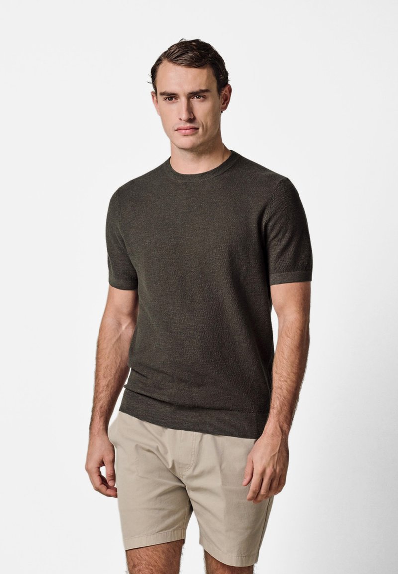 Homme portant un pull en maille à manches courtes vert foncé et un short beige arrivant au genou, debout devant un fond blanc uni.