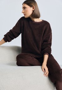 Brūna kažokāda atpūtas apģērbu komplekts ar brīvu piegriezumu, kas ietver ribotās aproces un apaļu kakla izgriezumu, novietots uz tekstūras gaiši pelēku virsmas.