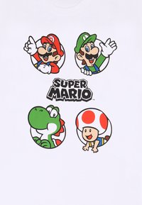 Bílý tričko s čtyřmi kulatými grafickými potisky: Mario, Luigi, Yoshi a Toad, s textem "Super Mario" v černé barvě pod obrázky.