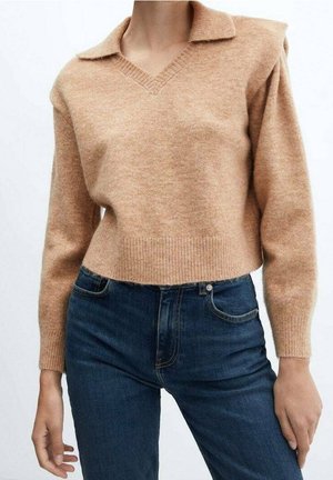 Person iført en beige langærmet striktrøje med V-hals og mørkeblå højtaljede jeans mod en ensfarvet baggrund.