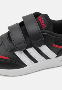 Zapatos para niños de color negro de material sintético con dos tiras de Velcro, franjas blancas en el lado y un acento rojo en el talón. Presentan un acabado texturizado.