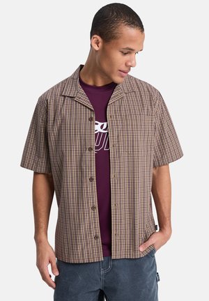 Kortermet, kragekjorte i brun og beige rutete mønster, med knappelukking foran og brystlomme, brukt over en lilla T-skjorte.