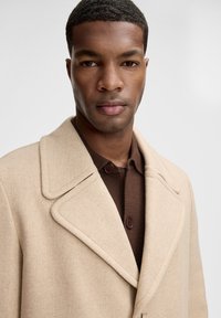 Cappotto beige in misto lana con ampio colletto a revers e chiusura a bottoni, indossato sopra una maglia lavorata marrone scuro con bottoni marroni.