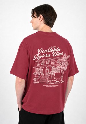 Mann trägt ein weinrotes T-Shirt mit weißer Skizze eines Gebäudes, Palmen und dem Text "Courtside Riviera Club" auf dem Rücken.